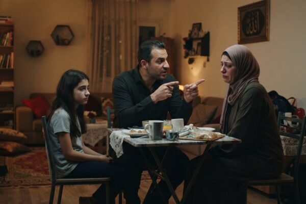 مشهد واقعي سينمائي لزوجين يجلسان في مساحة منزلية واحدة لكن يفصل بينهما توتر وصمت واضح، مع إضاءة هادئة وتكوين يعكس المسافة العاطفية بينهما، دون مبالغة أو عناصر رمزية غير واقعية